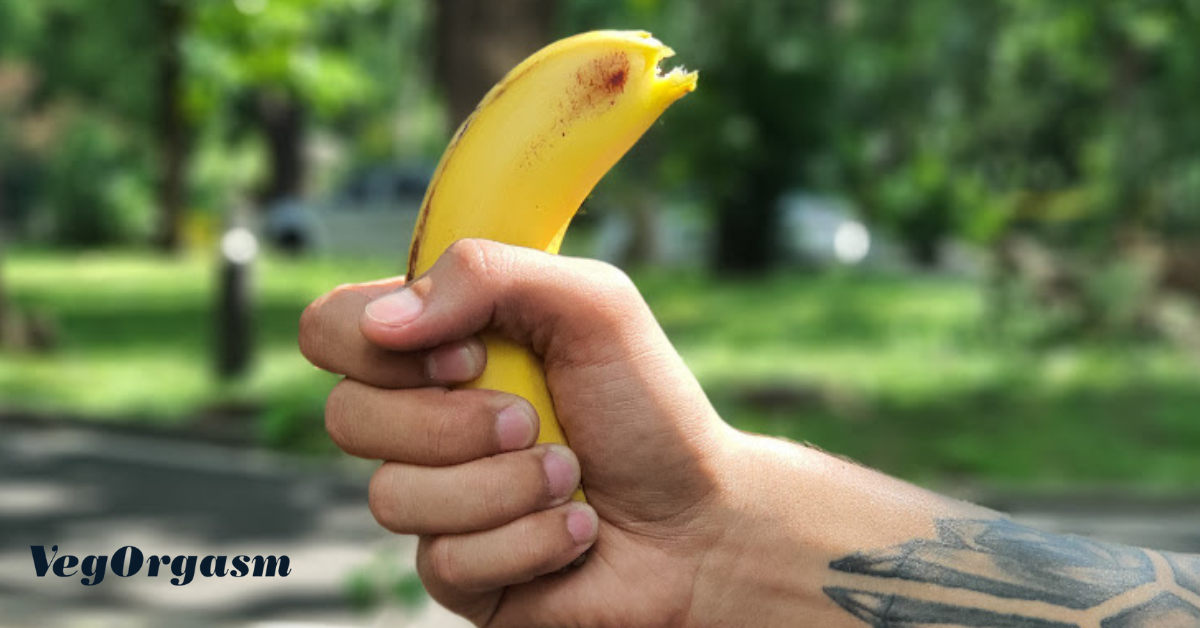 A close-up, horizontal image shows a person's hand with a tattoo on the forearm. The person is holding a half-peeled, slightly bruised banana in a tight grip. The background is a blurry park with green trees. A logo in the bottom left corner reads "VegOrgasam".