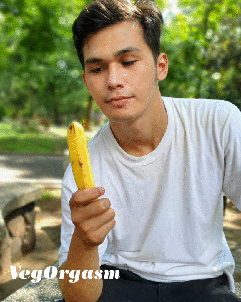 A young man with short dark hair and a white t-shirt looks at a slightly ripe banana he's holding. He is sitting outdoors on a stone bench with green trees and a path in the blurred background.
