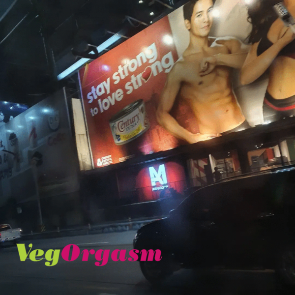 A night-time street view showing a large billboard illuminated above a building. The billboard features a shirtless alden richards with text that reads "stay strong to love strong" and prominently displays a can of "Century Tuna." Below the billboard, a "M AS GROUP" sign is visible. A dark car is in the foreground on the street, and a green and pink "VegOrgasm" logo is in the bottom left corner of the image.