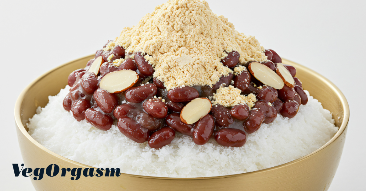 A wide-angled, eye-level shot shows a bowl of a popular Korean dessert called bingsu. The bingsu is set against a plain white background, making it the central focus. It is served in a shiny, golden-colored metal bowl. The bottom layer of the dessert consists of a mound of fine, snow-like shaved ice. On top of the shaved ice is a generous portion of sweet, cooked red beans, known as pat. The red beans are a deep reddish-brown color and have a glossy sheen. Mixed in with the red beans are several thin, oval-shaped slices of almonds. The entire dessert is crowned with a large pile of light brown powder, which is in-jeolmi, a Korean roasted soybean powder. The powder is loosely packed, giving it a fluffy appearance. In the lower-left corner, a black watermark with white lettering reads "VegOrgasm."