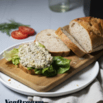 A top-down, angled shot of a vegan tuna salad sandwich on a small wooden cutting board. The open-faced sandwich is on a slice of whole-grain bread, topped with a piece of green lettuce and a generous scoop of chunky, light-colored tuna salad. Next to the sandwich, there are two more slices of bread and the end piece of the loaf. To the left, a couple of thin slices of red tomato and a sprig of dill are on the plate. A clear glass bottle and a black appliance are in the soft-focus background. The words "VegOrgasm" are written in a dark blue font in the bottom left corner of the image.