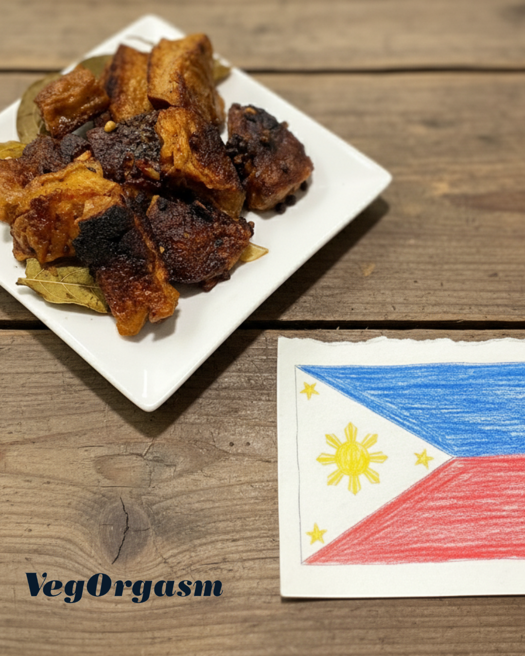 A plate of fried, crispy food adobo seitan with bay leaves sits on a rustic wooden table next to a childlike drawing of the Philippine flag.