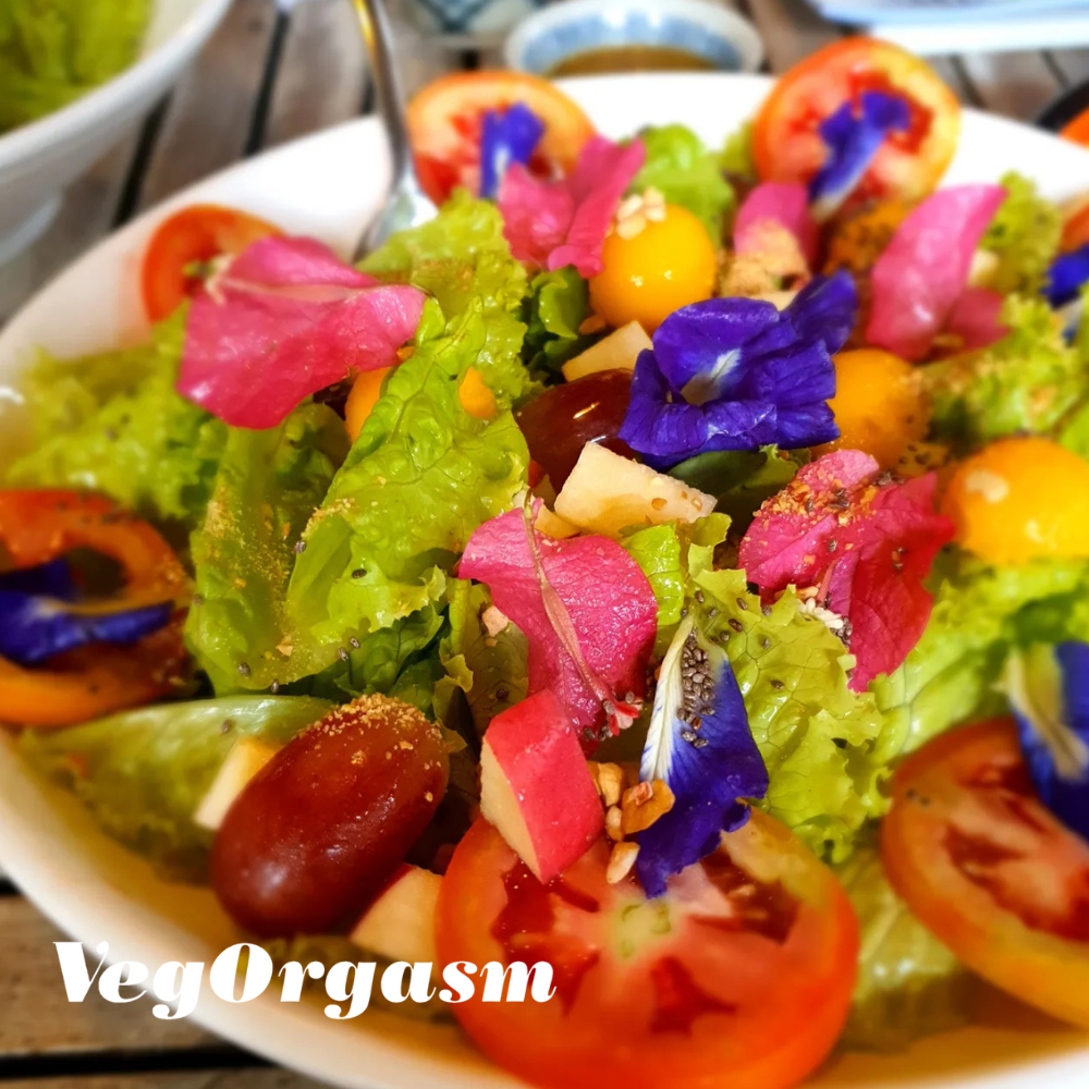 A vibrant salad is presented in a white bowl, filled with fresh green lettuce, sliced red tomatoes, and an assortment of colorful edible flowers in shades of pink, purple, and yellow. There are also whole red grapes, small yellow spherical fruits, and possibly some seeds sprinkled throughout. The background is softly blurred, showing what appears to be a dining setting. The word "VegOrgasm" is written in white, stylized font in the bottom left corner of the image.