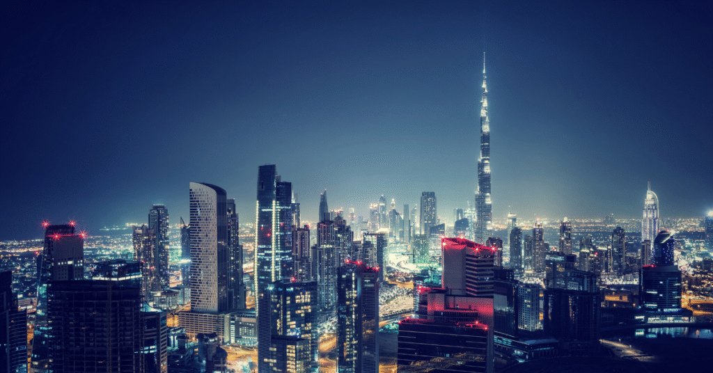 A high-angle, wide shot of the Dubai skyline at night, showcasing a dense cluster of illuminated skyscrapers under a dark blue sky. The iconic Burj Khalifa stands prominently in the background, its spire reaching far above the other buildings. Many of the buildings are lit with various colors, primarily white and blue, with some featuring red accents. The city lights stretch into the distance, suggesting a vast urban landscape. There's a subtle vintage or filmic quality to the image, with slightly desaturated colors and a hint of grain.