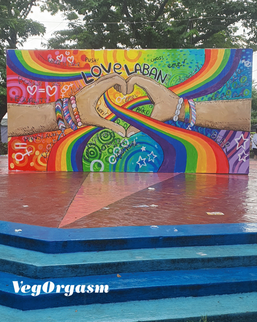 The fight for the SOGIE bill. A brightly colored, horizontal mural is displayed outdoors on what appears to be a stage or platform with blue steps leading up to it. The mural is a celebration of LGBTQ+ pride and solidarity.

The central image features two pairs of hands with light skin tones. The hands meet in the middle to form a heart shape with their thumbs and index fingers. They are adorned with colorful bracelets and beads. A vibrant, arched rainbow flows from the left side of the mural, passing through and beneath the hands, and continuing to the right side.  

The background is filled with a variety of colorful and diverse patterns, including hearts, stars, spirals, and scribbles. There are also numerous words and phrases written across the mural. Prominent text includes "LOVE LABAN" at the top center, and other phrases like "Love is Love," "Push!," "Equality," and names or phrases written in different fonts and colors. The style is bold and folk-art inspired.

The setting is outdoors, with wet ground suggesting it may have recently rained. Large trees are visible behind the mural. The "VegOrgasm" logo is visible in the bottom left corner of the image.