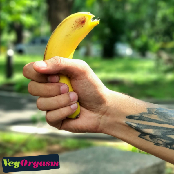 A low-angle, eye-level shot shows a man's right hand tightly gripping a partially peeled yellow banana. The fingers are curled around the banana, with the thumb visible on top. The man's forearm, which has a prominent tattoo of geometric, shard-like shapes, is also visible. The hand is positioned against a soft-focus background of green trees and grass, likely in a park or backyard. In the bottom left corner, a logo with the text "VegORGASM" is visible in a white, stylized font on a black and green background. The image has a slightly yellow tint and appears to be well-lit.