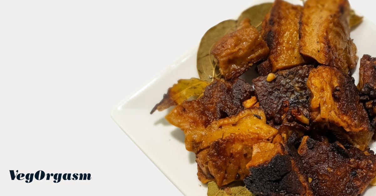 A close-up shot of a white square plate filled adobo - featuring crispy, golden-brown pieces of seitan. Some dried bay leaves are scattered amongst the food. The background is plain white. In the bottom left corner, the text "VegOrgasm" is visible in a stylish, dark font.