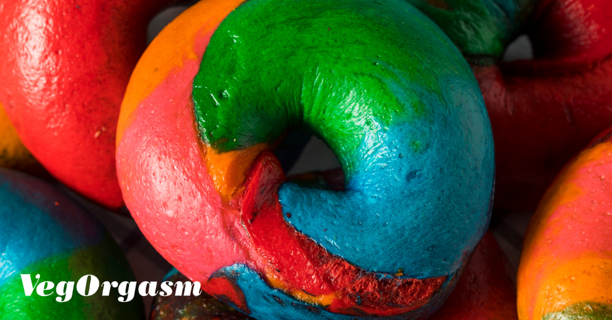 A very close-up, horizontal photograph focuses on a single, vibrant rainbow bagel, with others blurred in the background. The central bagel is swirled with bright, saturated colors—including green, blue, red, pink, and orange—which are clearly defined and glossy. The texture of the bagel's surface is smooth. Other rainbow bagels are partially visible on the left and right, out of focus. The "VegOrgasm" logo is visible in the bottom left corner.