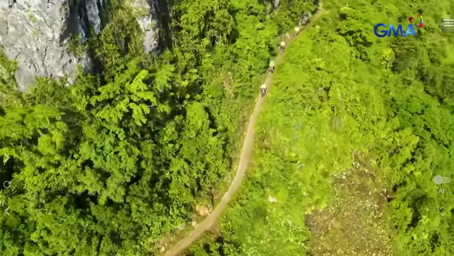 Aerial view of farmers carrying heavy sacks of produce on a narrow, unpaved, winding dirt path surrounded by dense green jungle and a rocky cliff face, illustrating the challenging transport conditions in Jessica Soho's farm-to-market road investigative exposé.